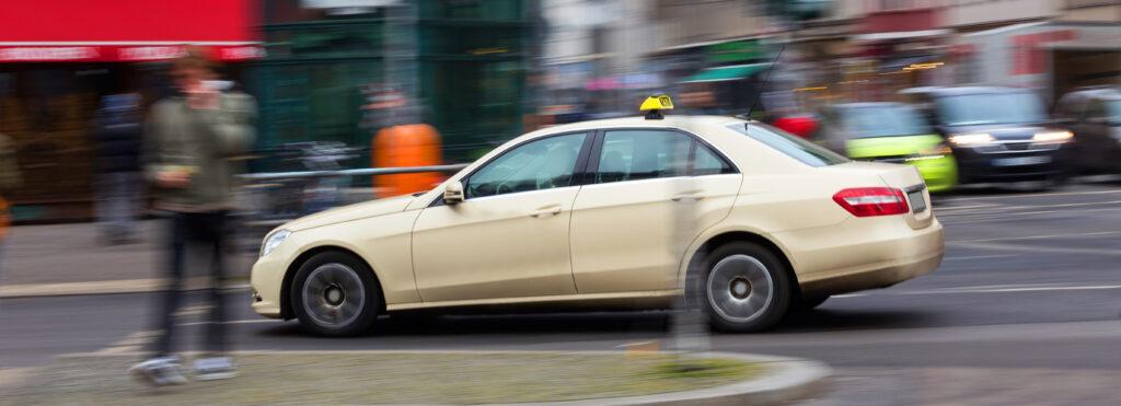 Ein Taxi fährt durch die Stadt, während ein Passant vorbeigeht – Alltag im Taxifahrer Job.