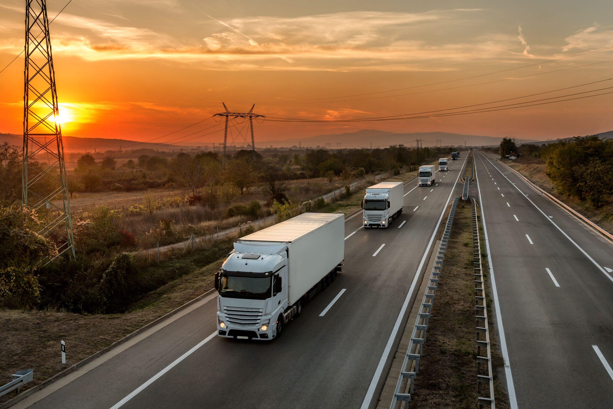 Konvoi blauer Lastwagen auf einer Landstraße unter einem atemberaubenden orangefarbenen Sonnenuntergangshimmel. Autobahntransport mit weißen LKW-Spuren in Karawane oder Konvoi